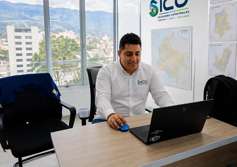 Profesional de SICO analizando información catastral en un portátil, con mapas de Colombia y el logotipo de la empresa al fondo